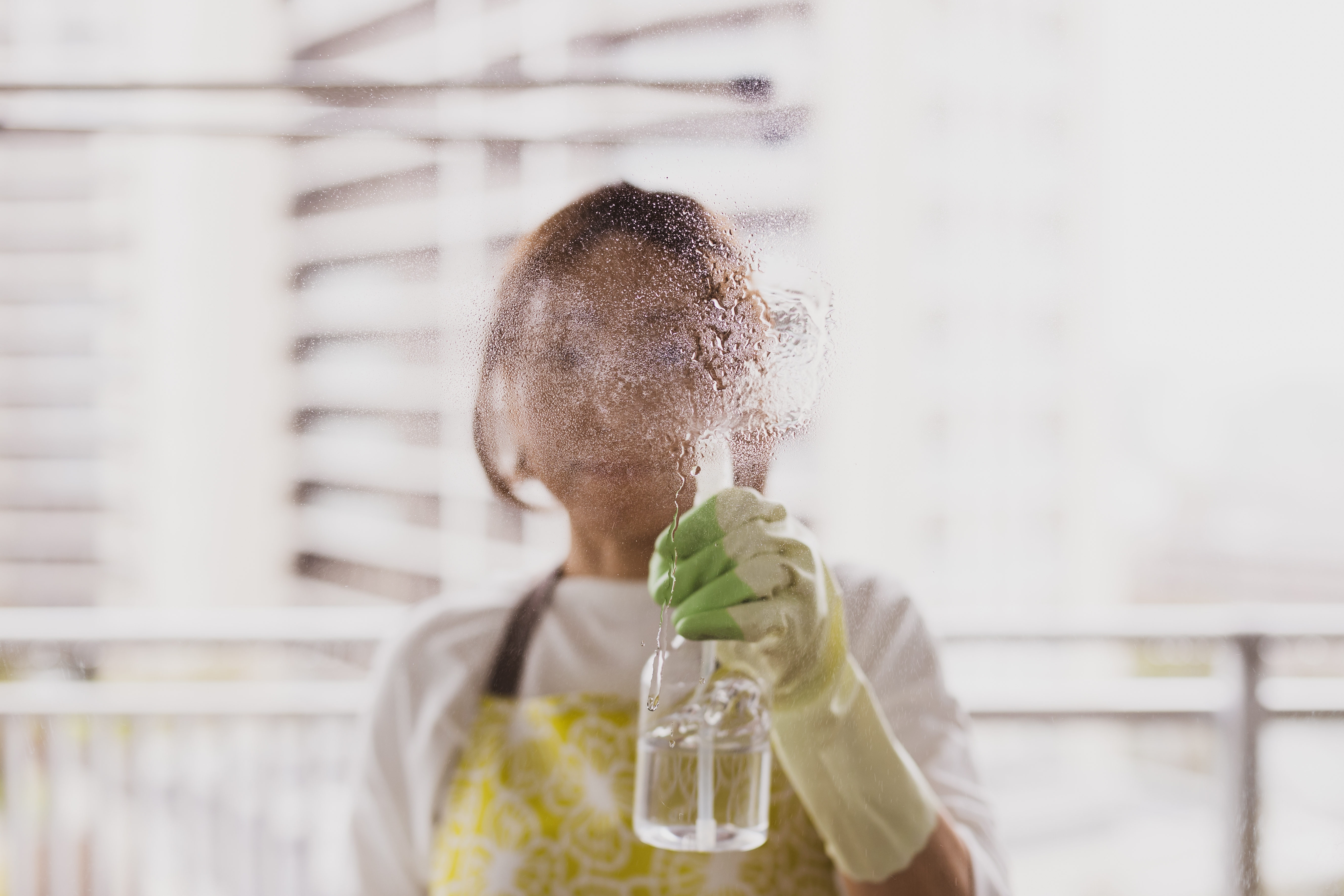 Person spraying face with mist from a bottle, standing on a balcony, wearing gloves and an apron