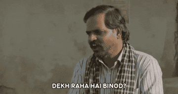 Man with a plaid scarf and striped shirt animatedly gestures while speaking Hindi text: &quot;Dekh Raha Hai Binod?&quot;