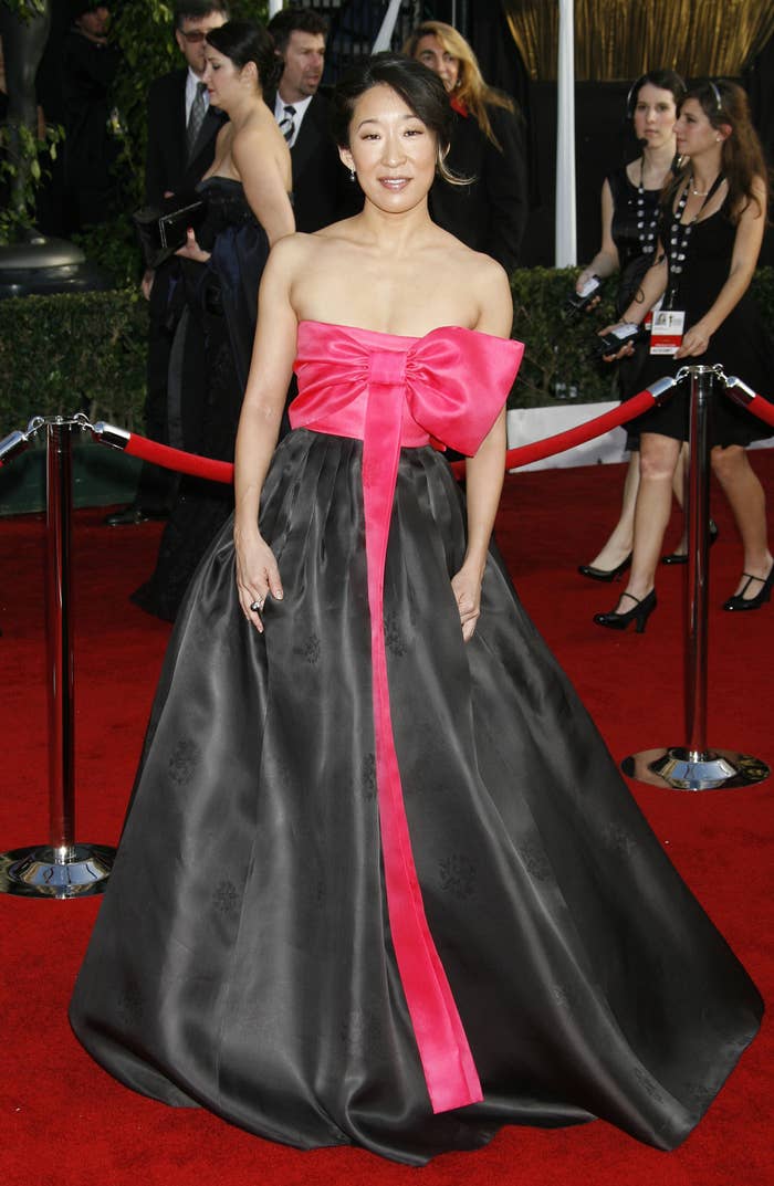 Person in a strapless gown with a large bow stands on a red carpet, surrounded by attendees and photographers