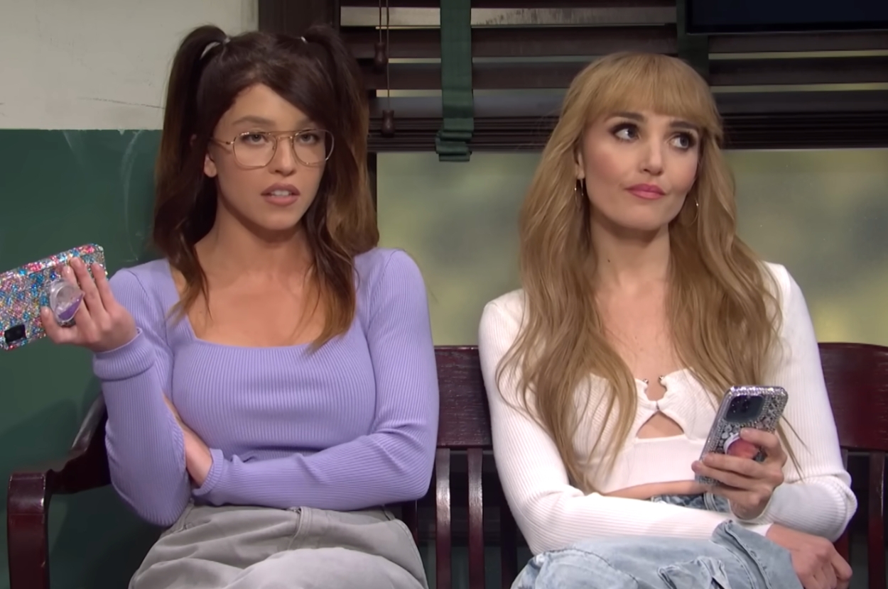 Two women sit on a bench, holding phones and wearing casual tops with jeans, engaged in a conversation