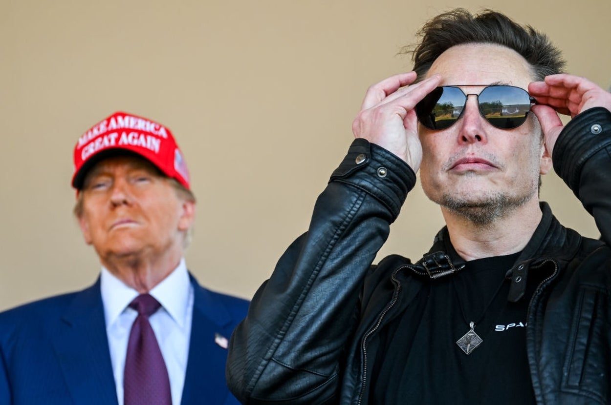 Two men standing; one in a suit with a red cap, the other in a leather jacket adjusting sunglasses