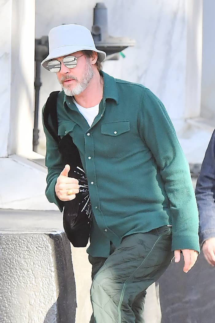 Brad Pitt in casual green shirt and pants, wearing a white bucket hat and sunglasses, holding a black bag, walking outdoors