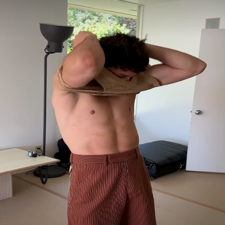 Person indoors putting on a knitted top, with striped pants in a casual setting. Standing near a lamp and an open door