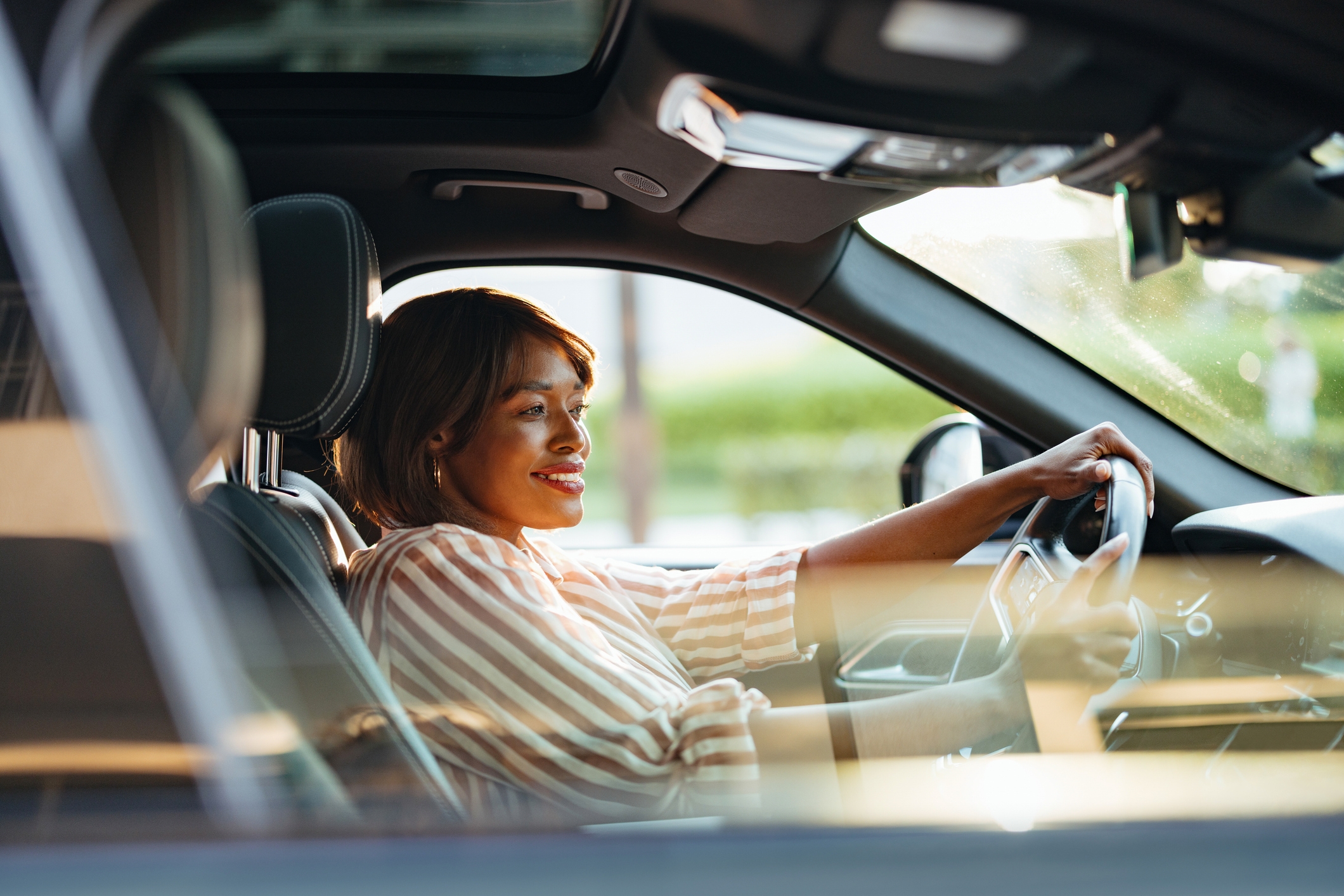 En smilende person sitter i en bil og holder rattet selvsikkert og har på seg en stripet skjorte
