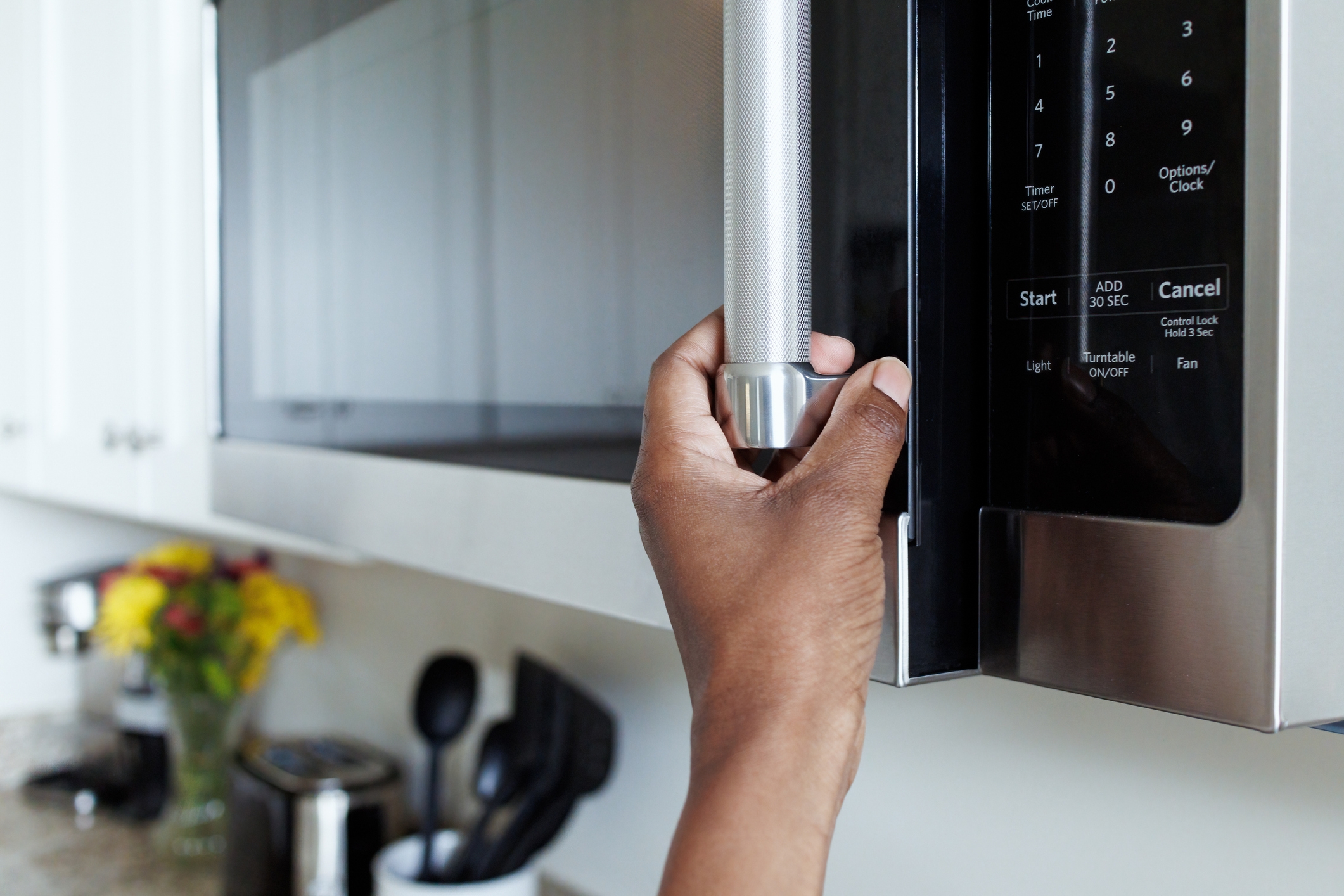 Hand opening a microwave door in a kitchen, with kitchen utensils and a vase of flowers in the background