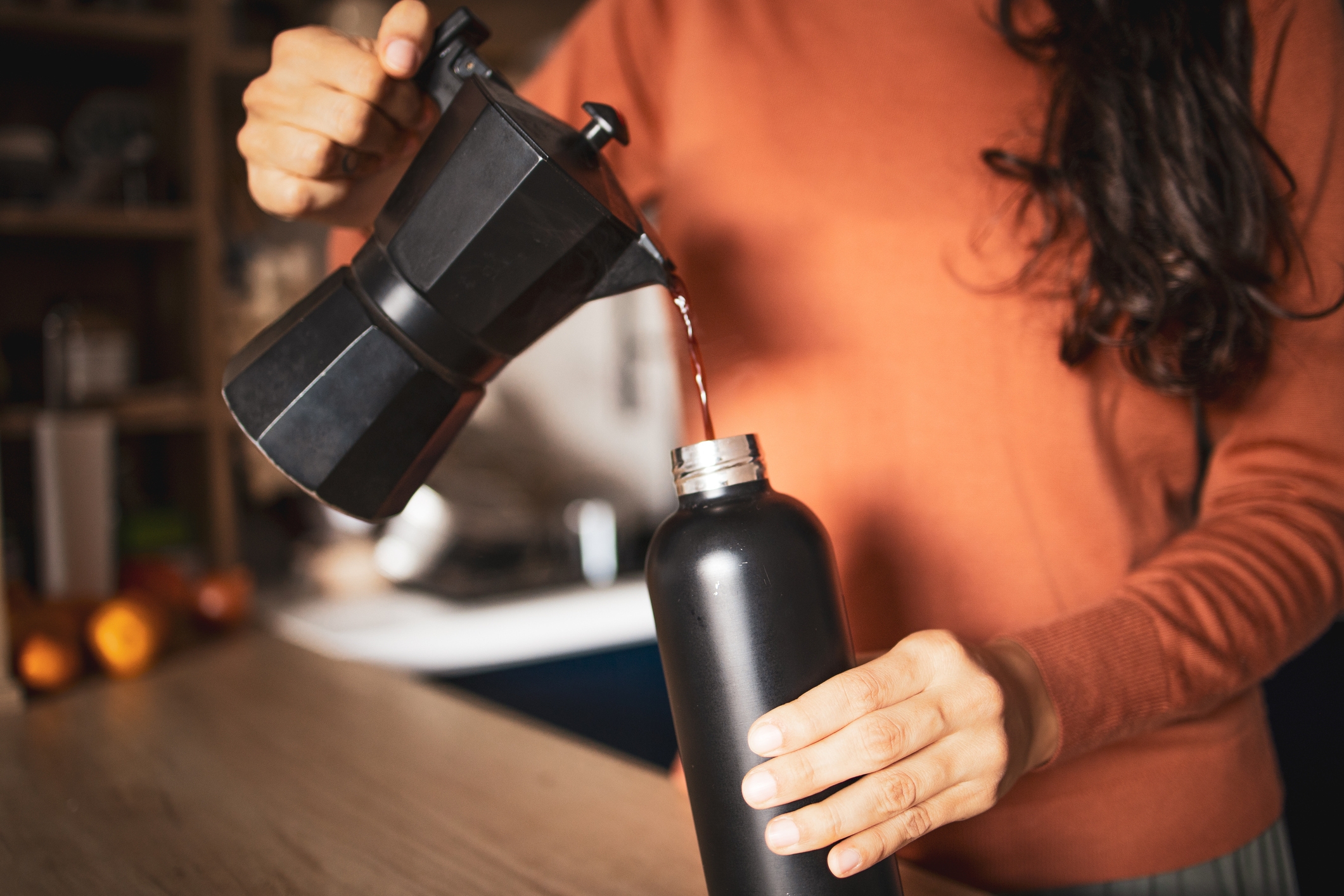 A person pours coffee from a moka pot into a black thermos, in a cozy kitchen setting