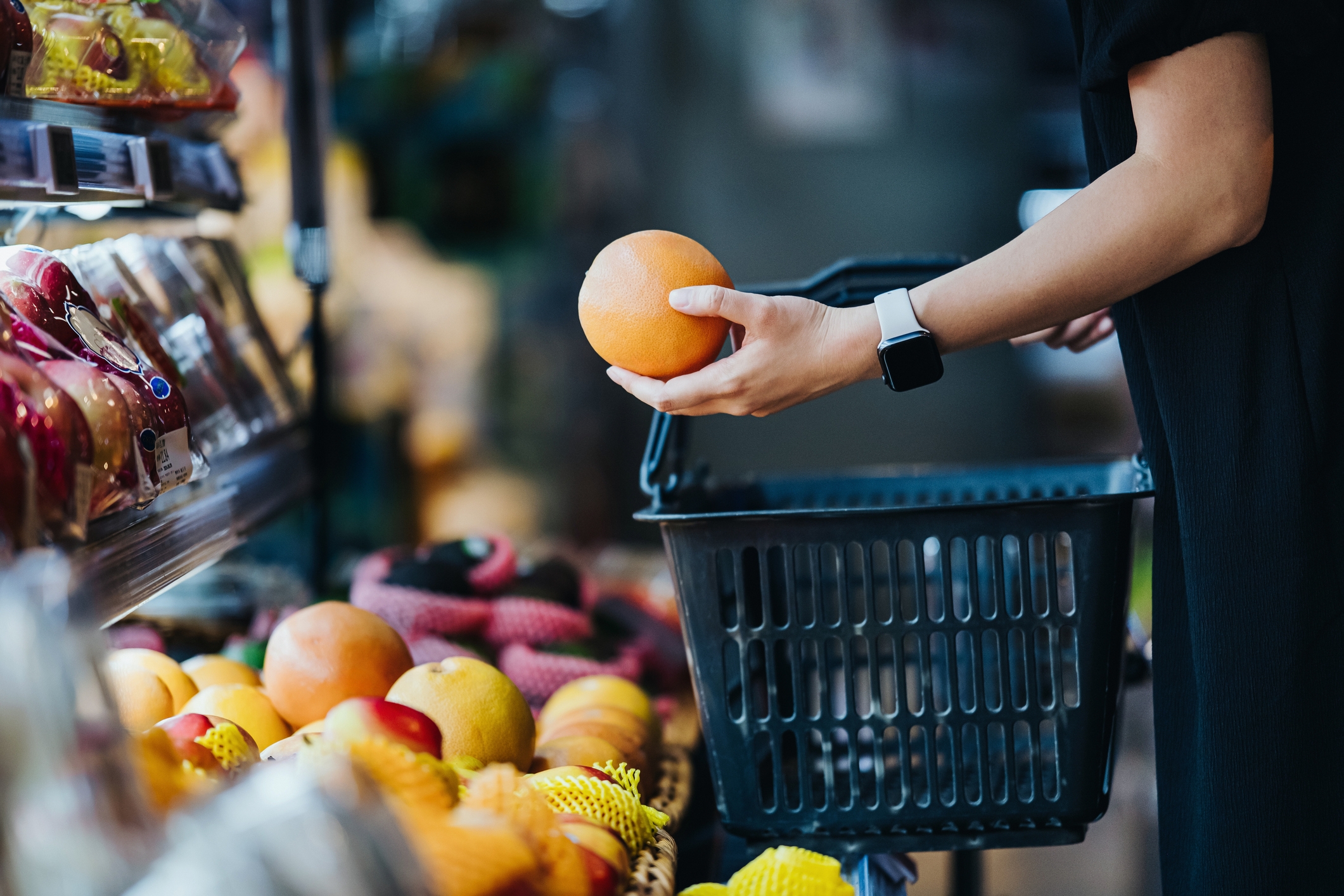 Person with smartwatch choosing an orange at a grocery store, holding a shopping basket surrounded by fresh produce