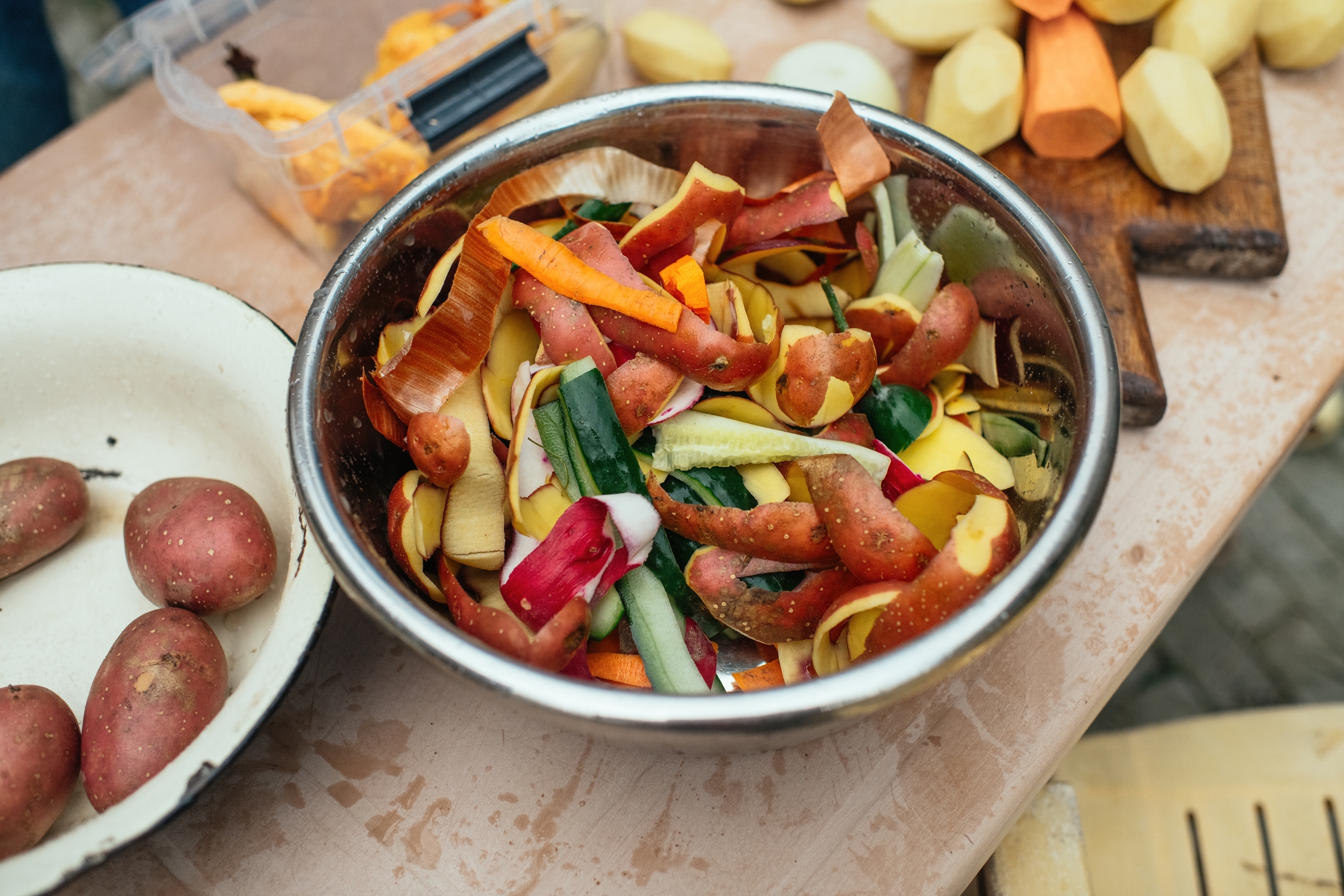 A bowl of assorted vegetable and potato peels on a table, surrounded by whole potatoes and a cutting board with chopped veggies