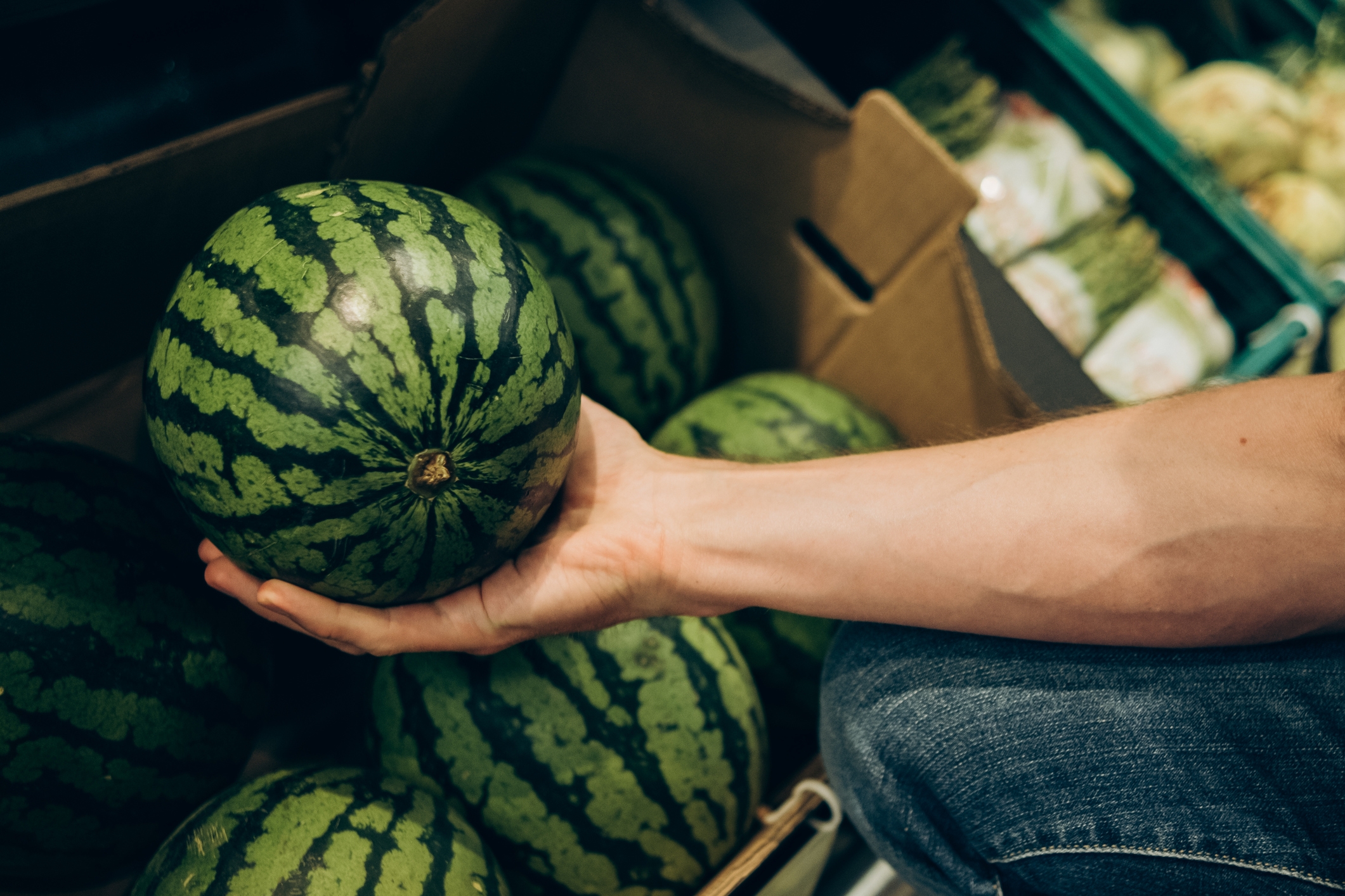 Person holding a watermelon in a grocery store, with other watermelons visible in a box