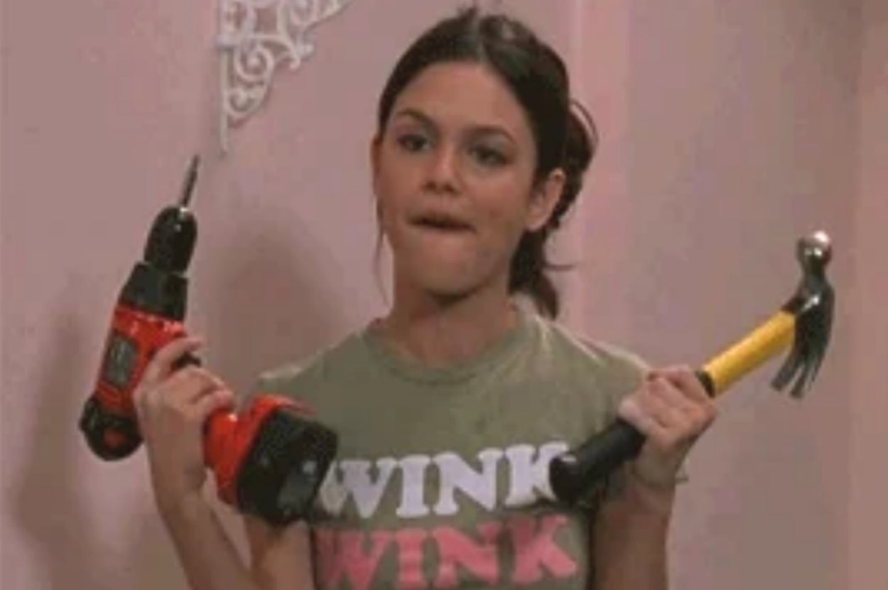 A woman playfully holds a drill and a hammer, wearing a t-shirt with text
