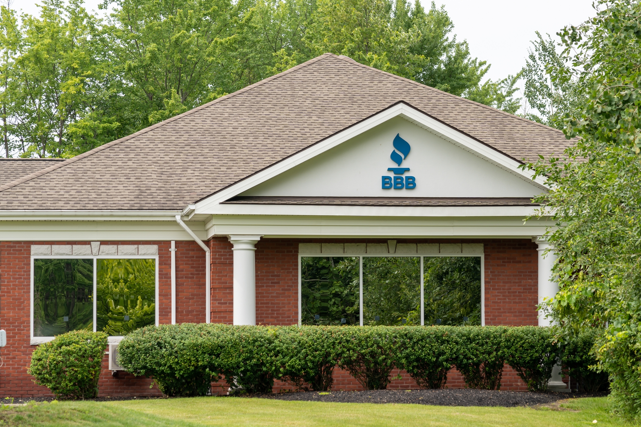 En murbygning med BBB-logoen på fasaden, omgitt av trær og velstelte busker