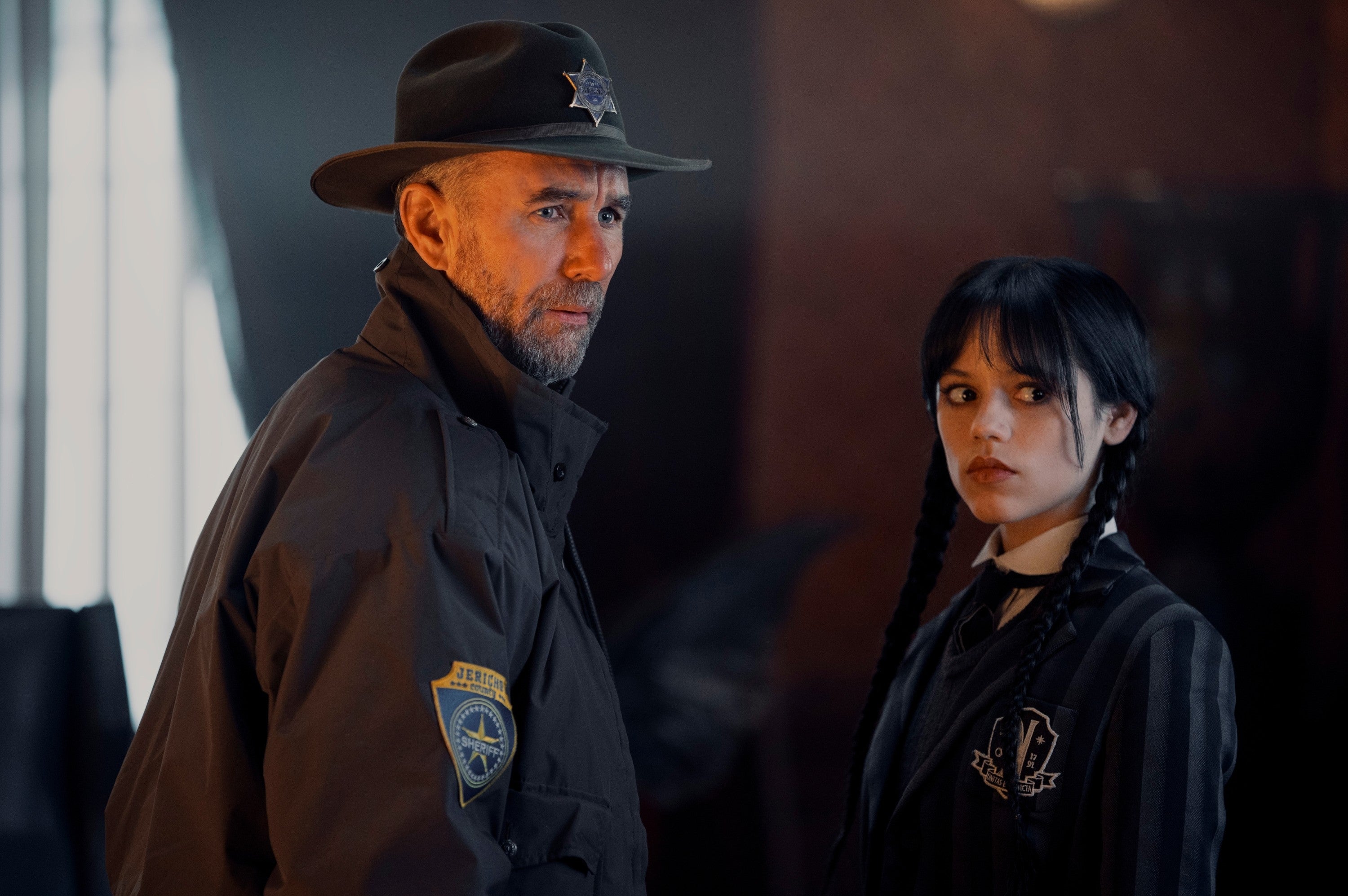 Two actors in a dramatic scene; one wears a sheriff's coat and hat, the other wears a striped blazer with long braids, looking serious