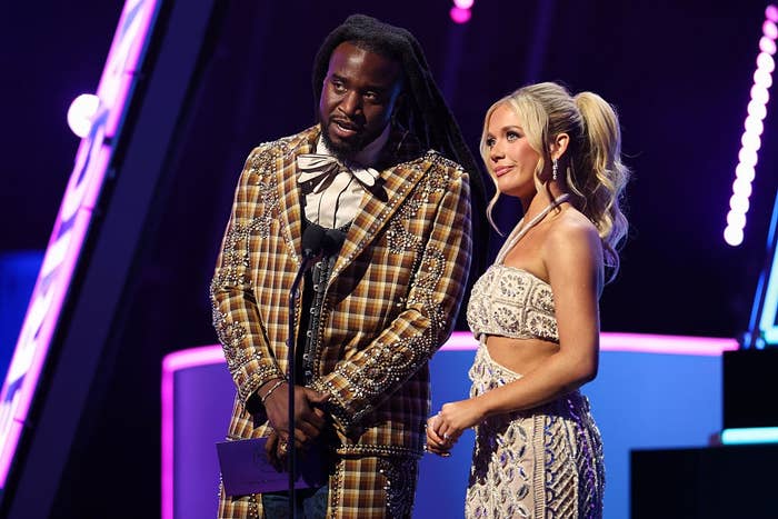 Two celebrities on stage. The man wears a patterned suit with embellishments, and the woman wears a strapless, embroidered gown. Both hold a microphone