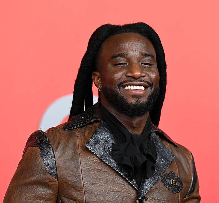 A person smiles on the red carpet wearing a textured brown leather jacket with a black ruffled shirt underneath