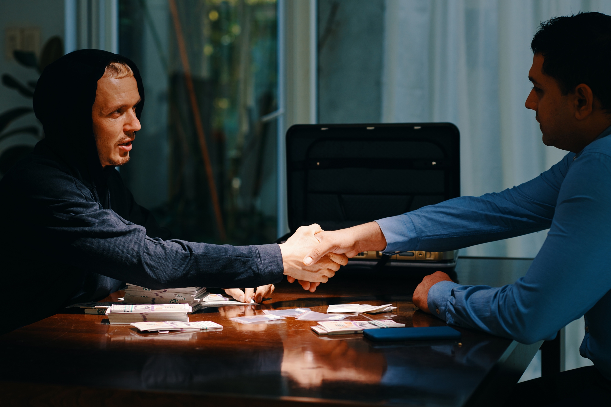 To personer ved et bord håndhilser over spredte materialer, som symboliserer enighet eller forhandling. En koffert er åpen i bakgrunnen