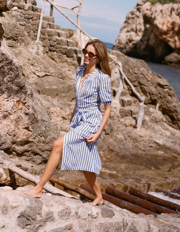 Person successful a striped formal walks barefoot connected rocky terrain by the sea. Wearing sunglasses, she exudes a casual, chic style