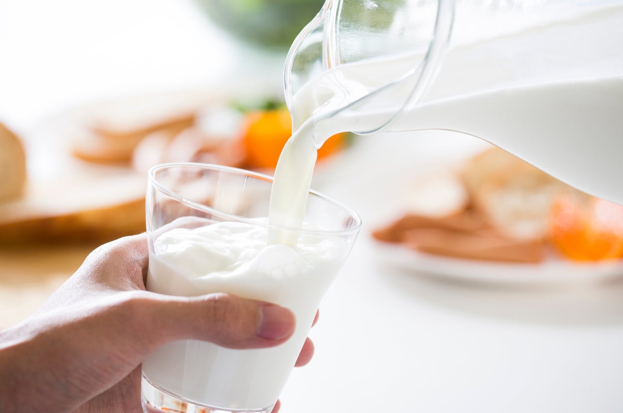 グラスに注がれる牛乳。背景にパンや果物がぼんやりと見える。