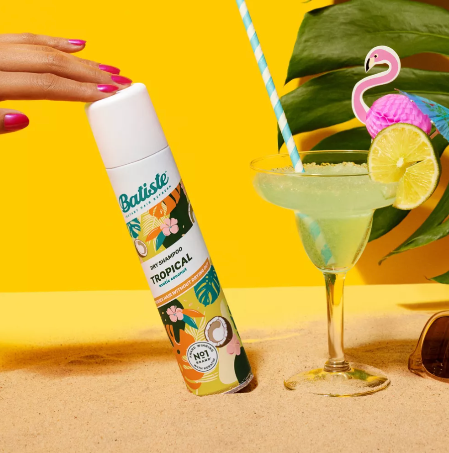 Batiste Tropical Dry Shampoo di sebelah minuman tropis dengan sedotan flamingo di atas pasir, memberikan getaran pantai