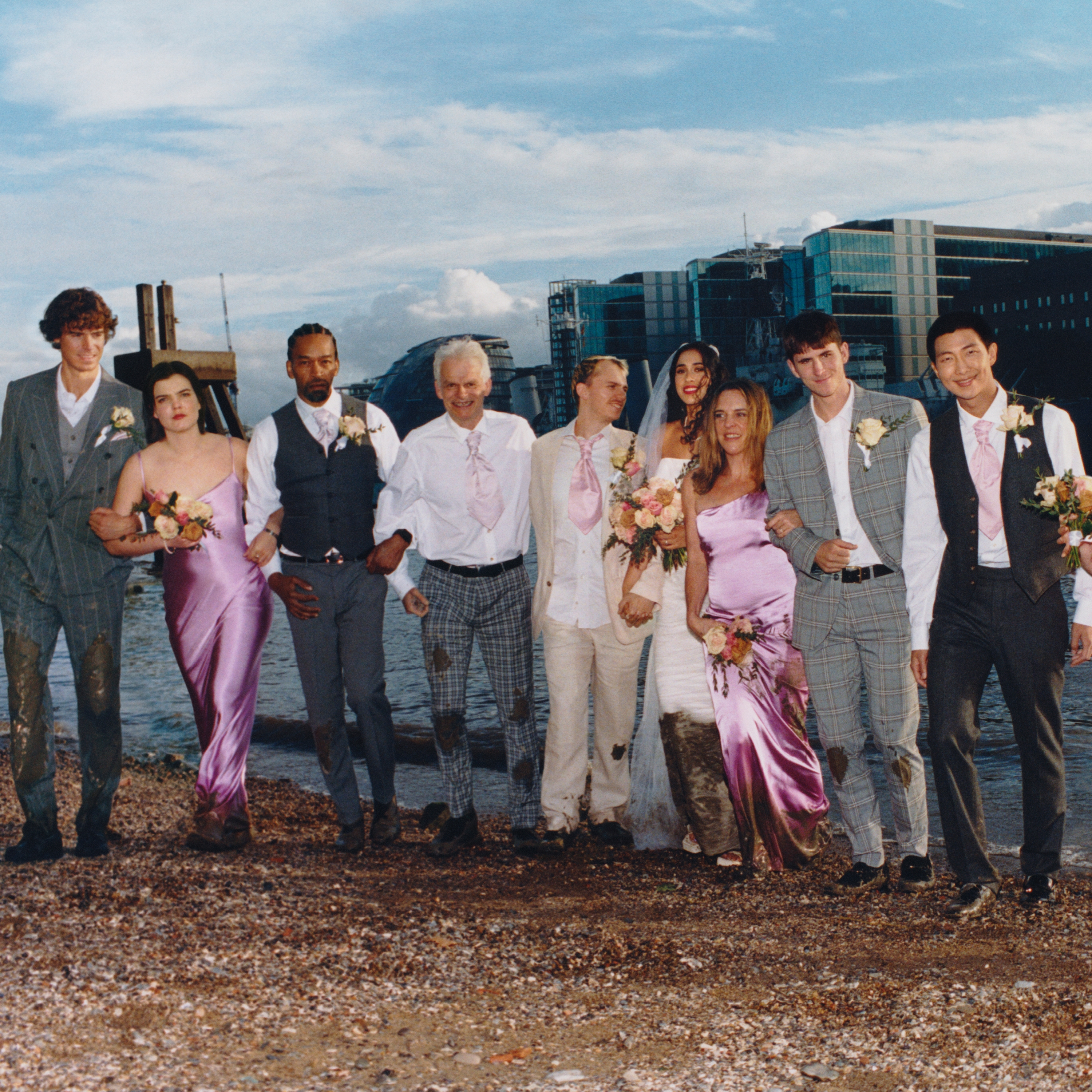 Group of people in formal wear, including suits and dresses, posing on a pebbled shore with buildings and water in the background