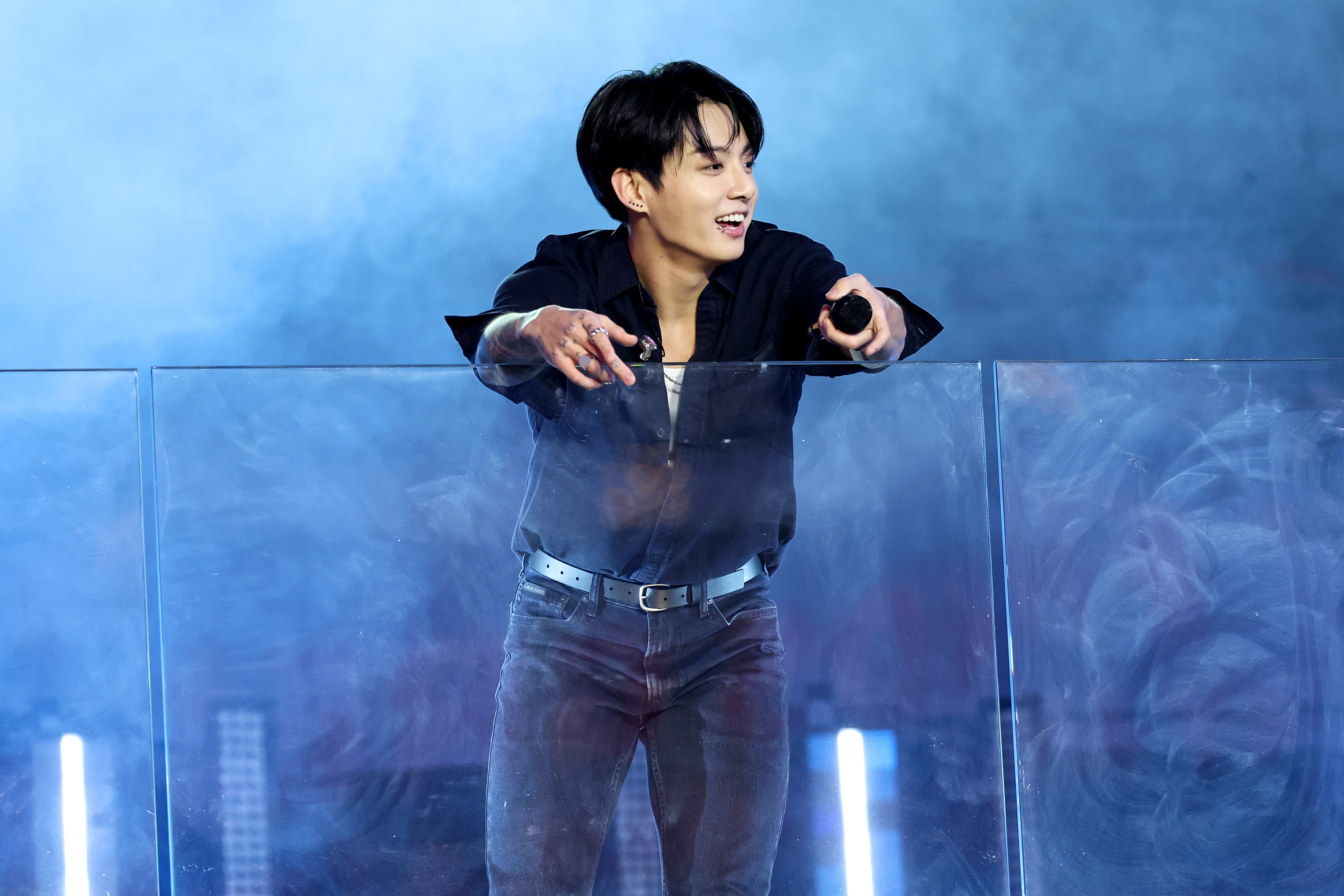 A person in a casual button-up shirt and jeans sings energetically on stage behind a clear barrier