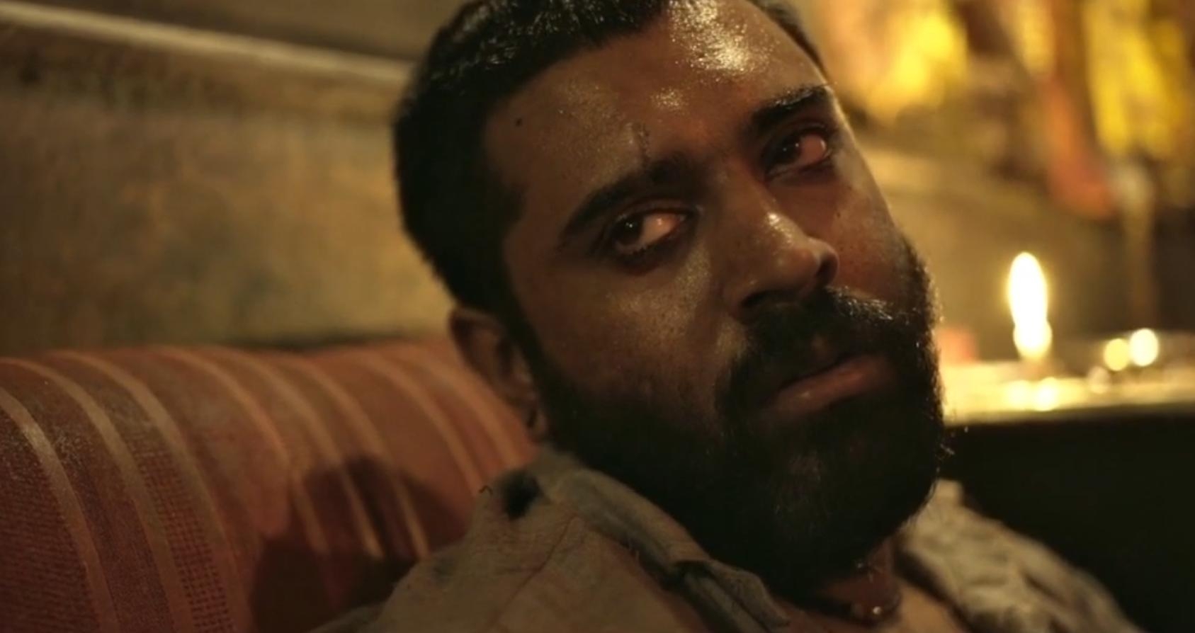 Person with a beard appears tired and sweaty, leaning against a striped surface in dim lighting