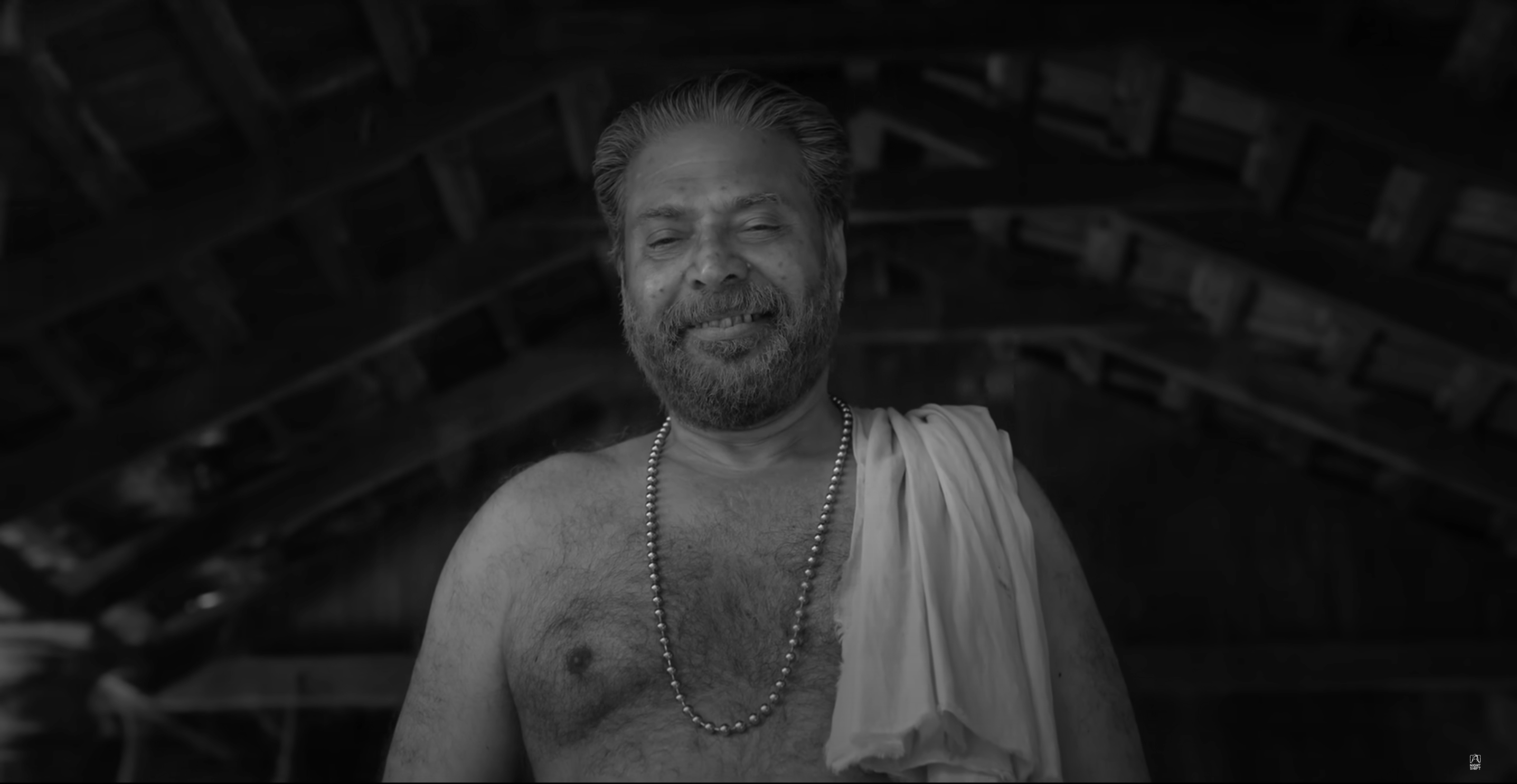 Smiling man wearing a beaded necklace and draped cloth stands under a wooden roof