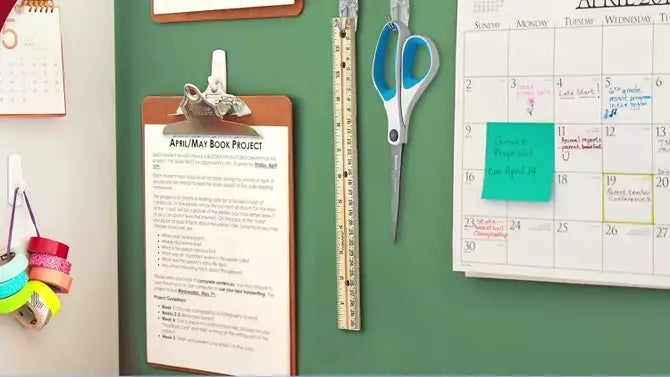 Kalender dan papan klip yang dipasang di dinding untuk proyek buku dengan selotip, gunting, dan penggaris di dekatnya. Kamar terang, ruang kerja terorganisir