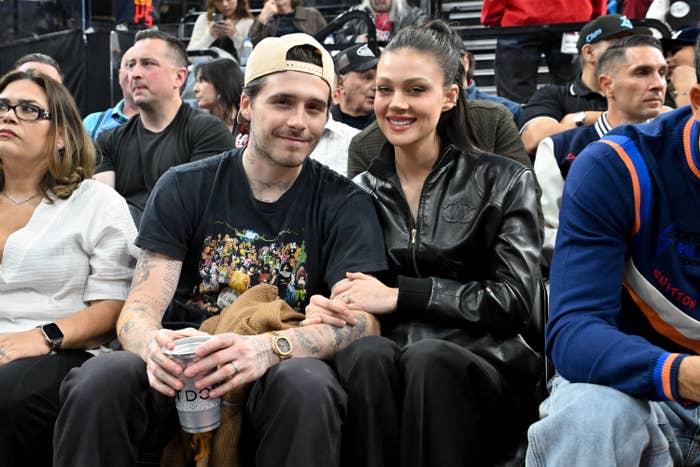 Two people sitting courtside at a sports event, smiling and holding hands. One wears a baseball cap and T-shirt; the other is in a leather jacket