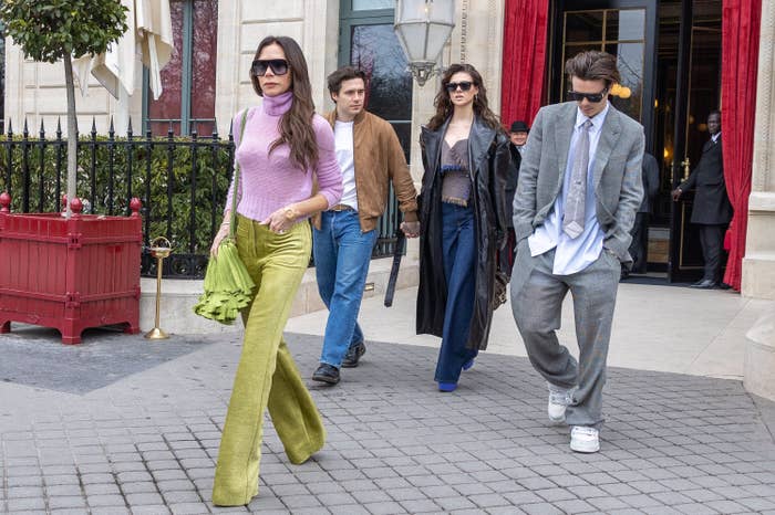 Four individuals in stylish outfits walk outside a building. One wears a turtleneck and flared pants; others wear casual and tailored clothing