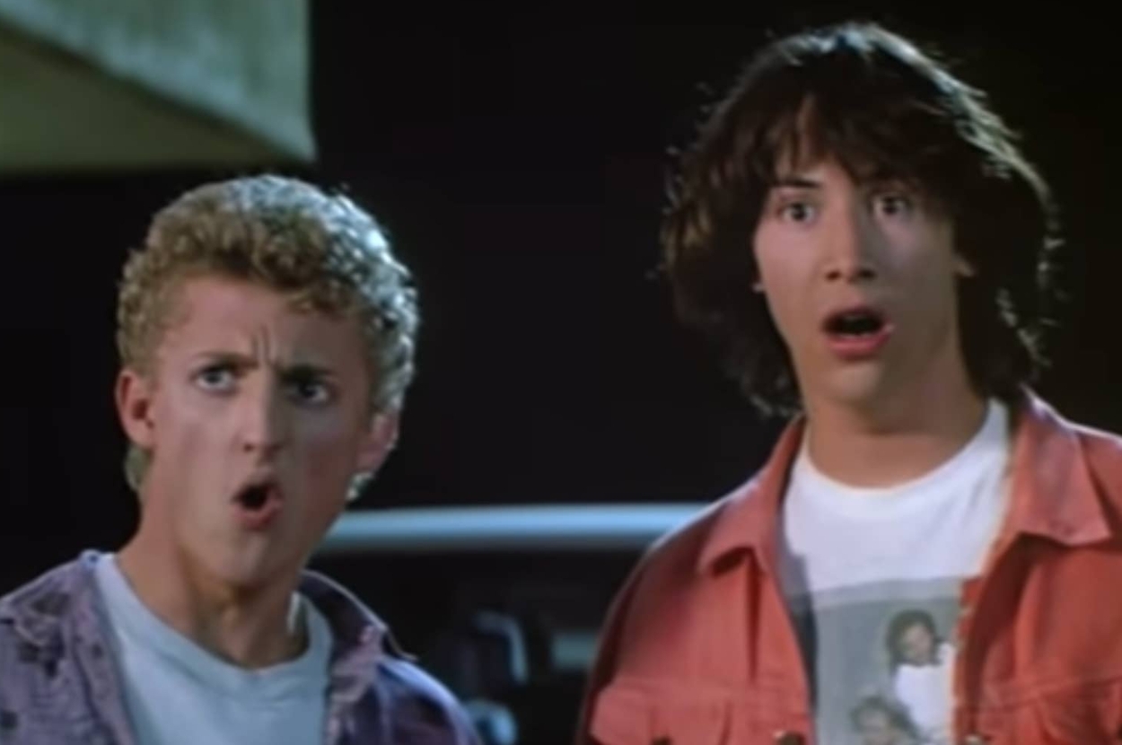 Two young men with surprised expressions, wearing casual 80s-style clothing, one in a denim jacket and the other in a T-shirt and open shirt