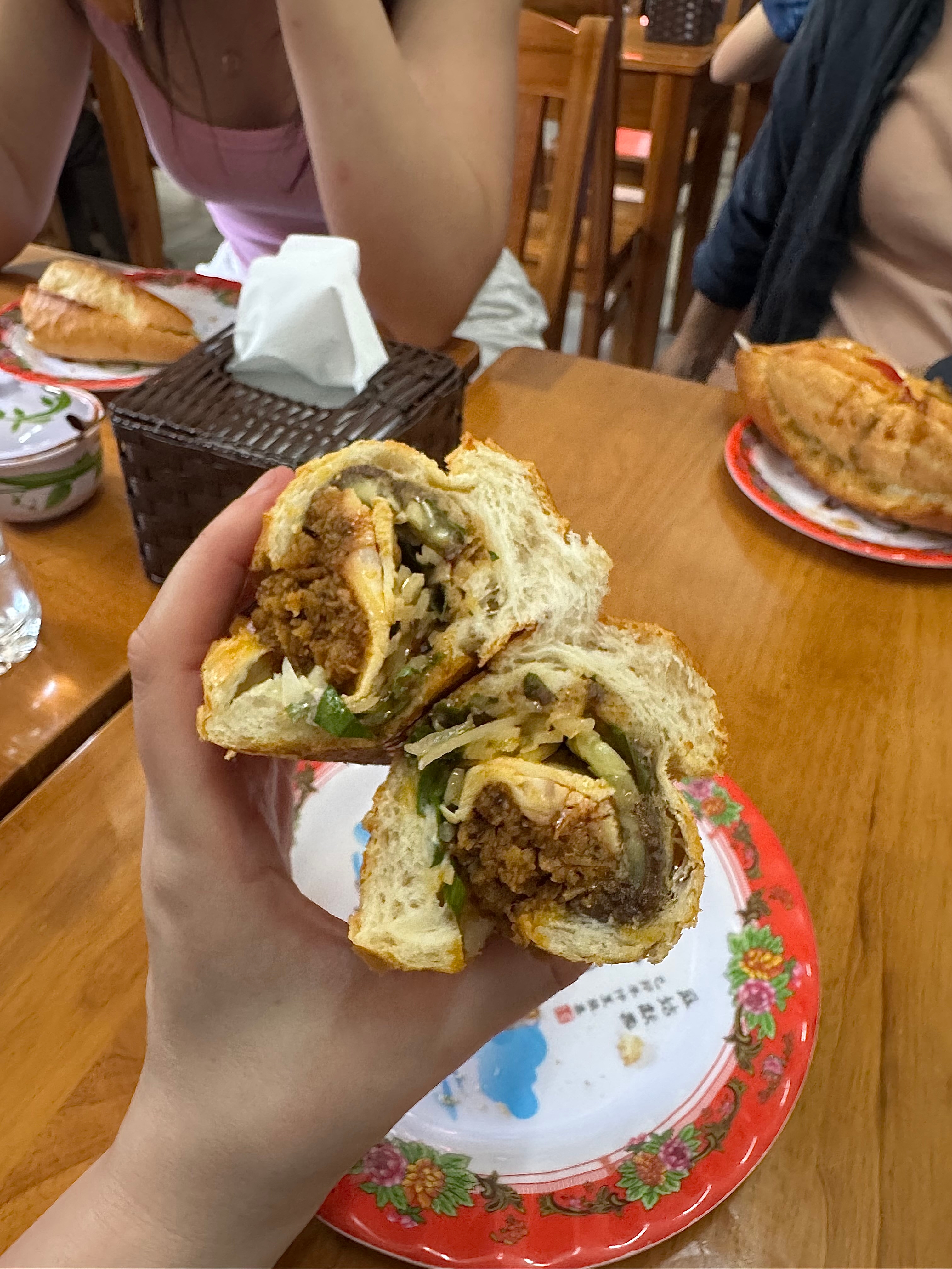 A hand holds a bánh mì sandwich filled with meat, vegetables, and herbs over a wooden table with people dining in the background