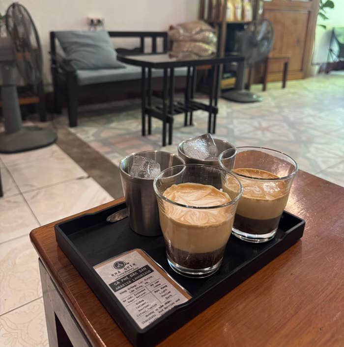 Three glasses of coffee served on a small tray, placed on a table in a cozy café setting with seating in the background