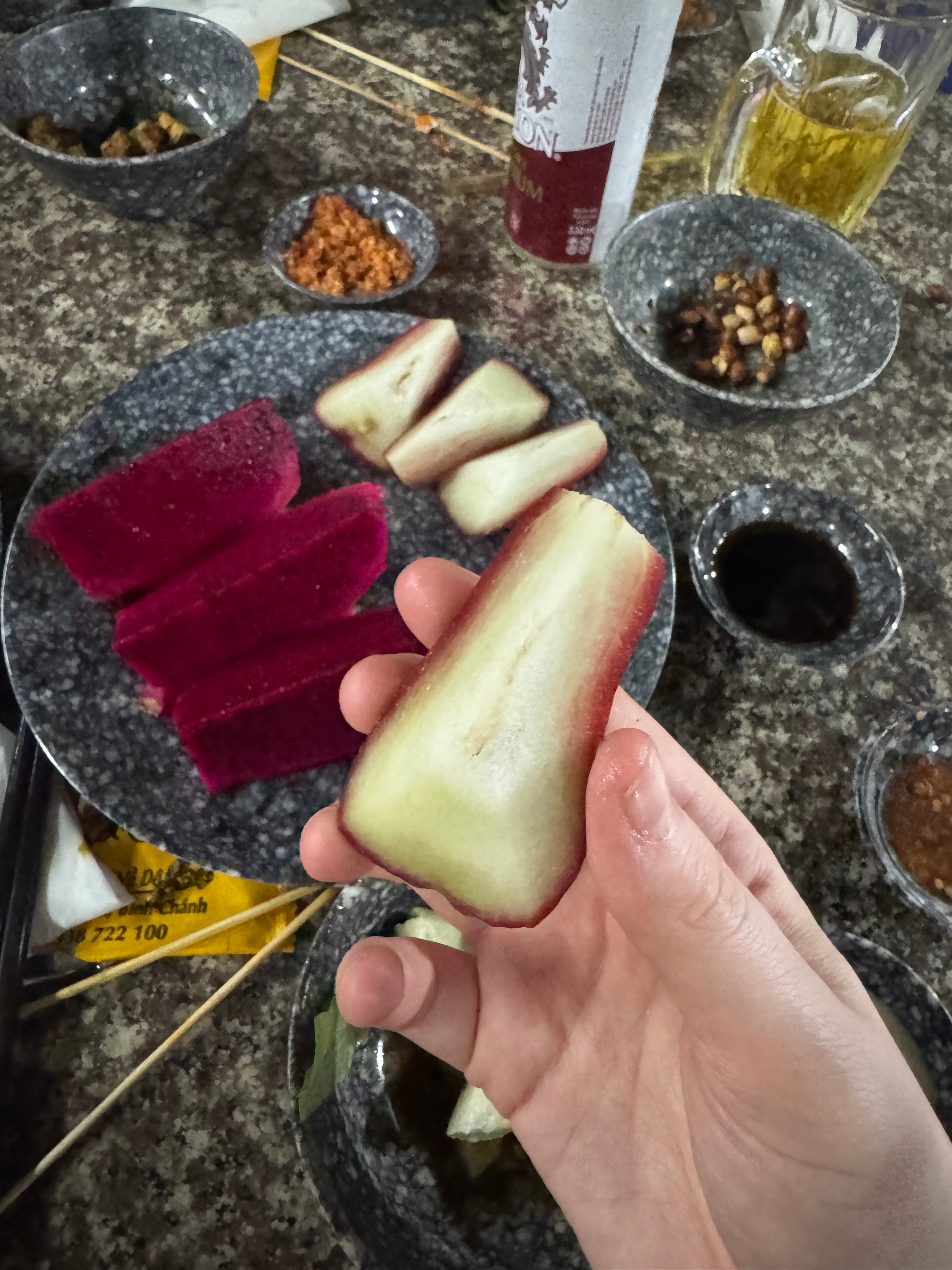A hand holds a white and red fruit slice over a table with various snacks, sauces, and a drink, suggesting a casual dining setting