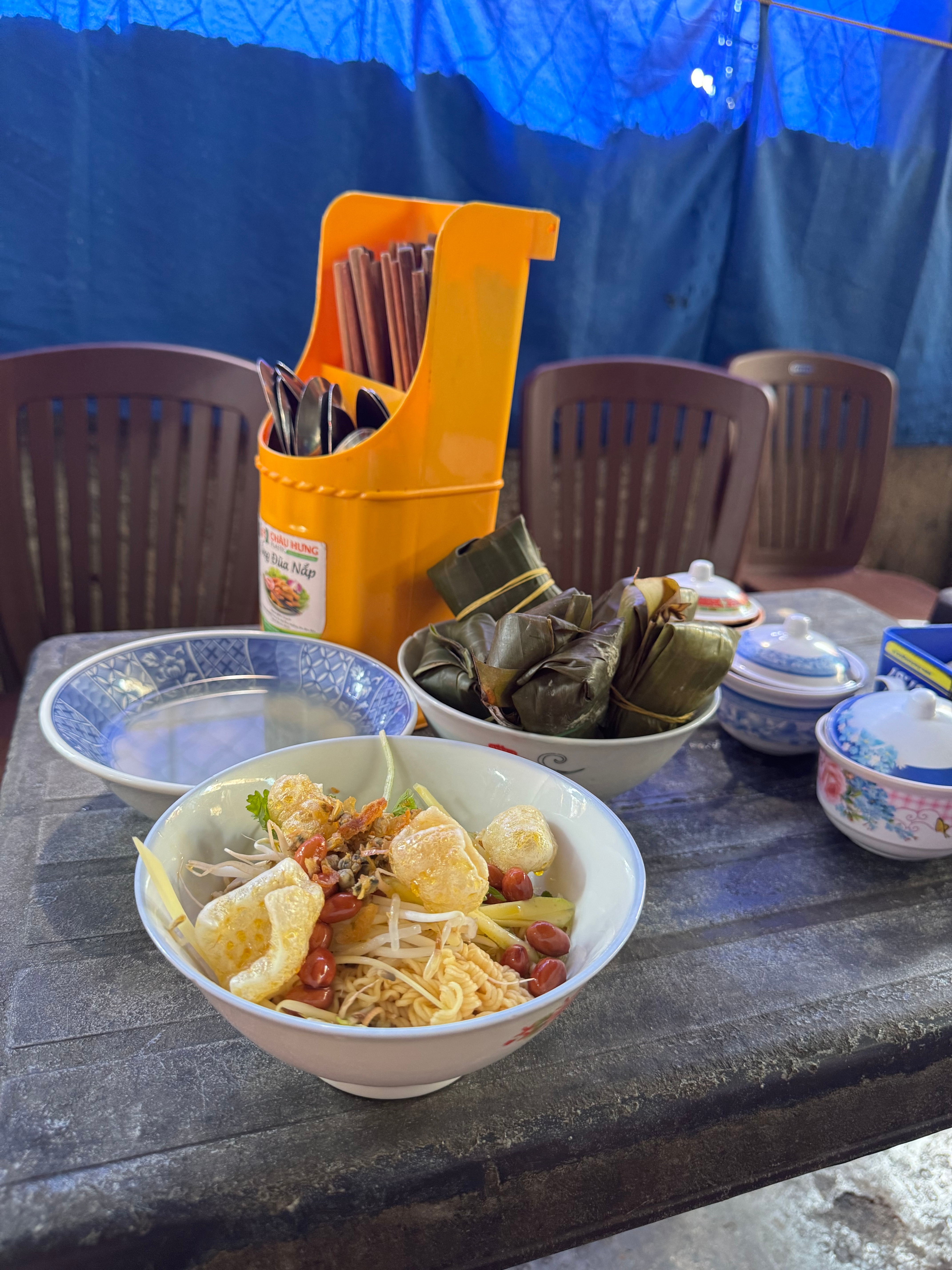 Bowl of noodles with toppings and a bowl of wrapped rice cakes on an outdoor table with utensils nearby