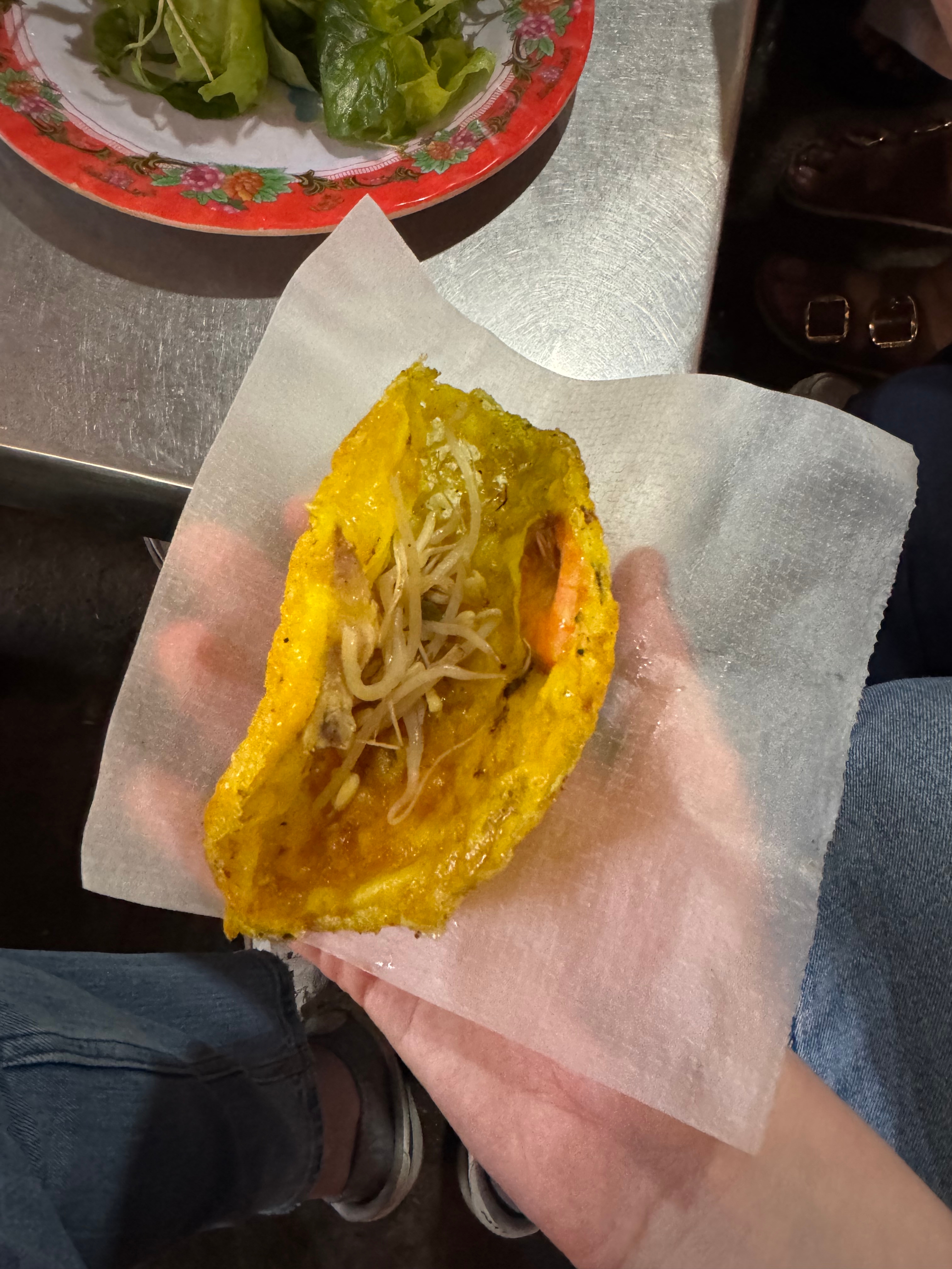 Person holding a folded cornbread with stir-fried vegetables, on paper, above a metal table with a plate of greens