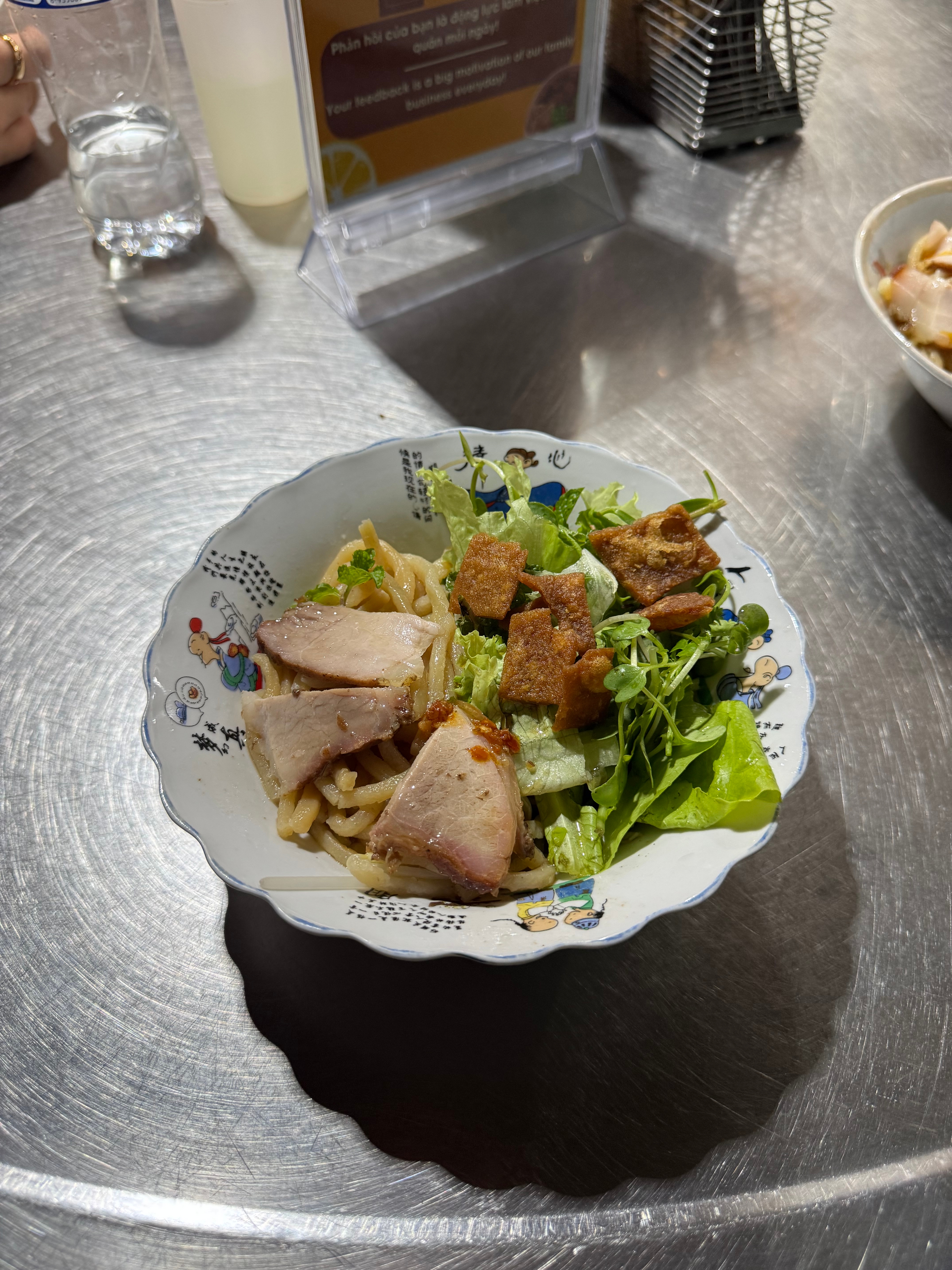 A bowl of noodles with slices of meat, greens, and croutons on a decorative plate, placed on a metal surface