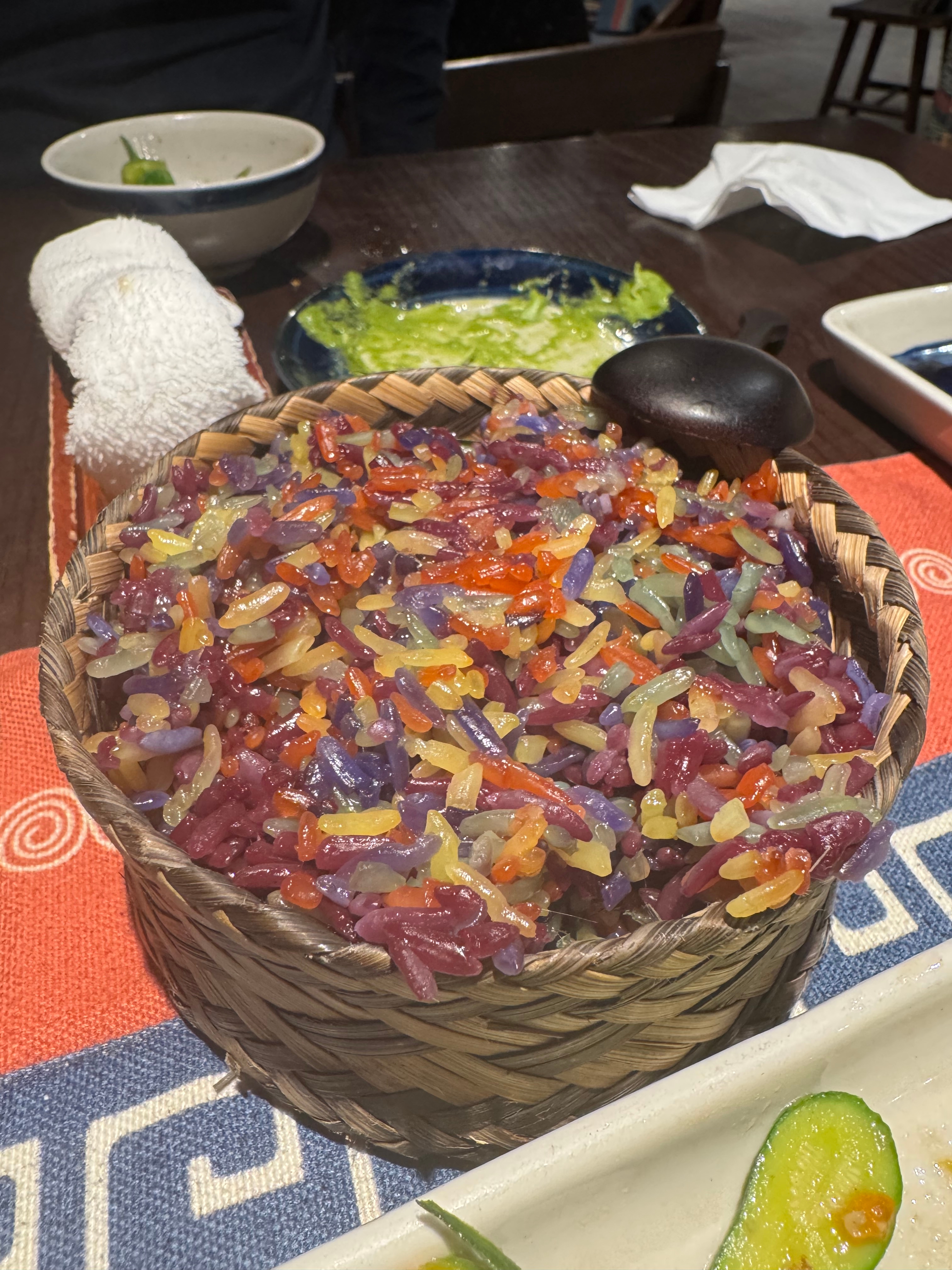 Basket of multicolored rice on a table set for a meal, served with a black spoon and white napkin in background