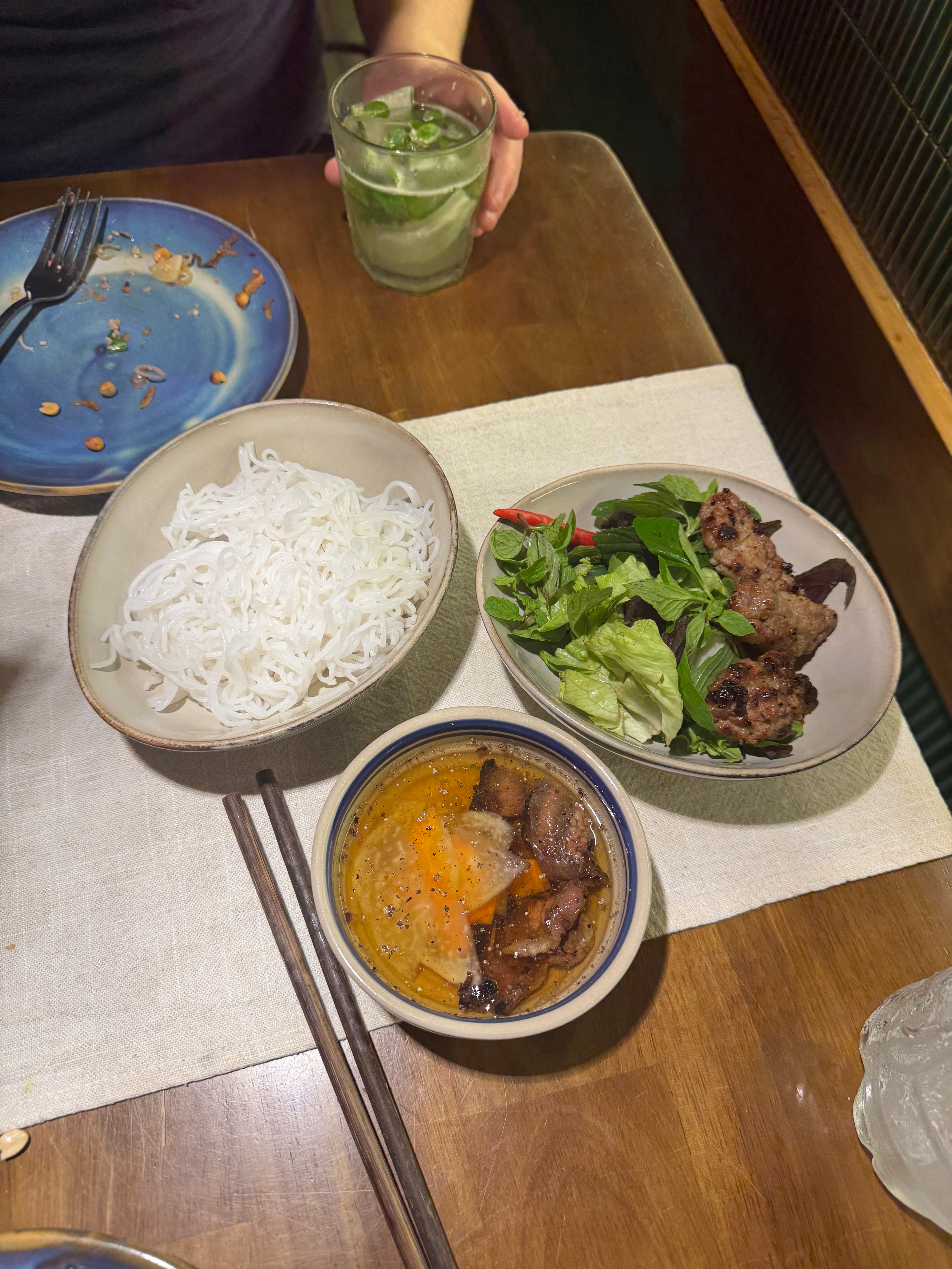 A table with Vietnamese noodle soup, grilled meat, fresh greens, and a drink. Chopsticks are placed beside the bowls
