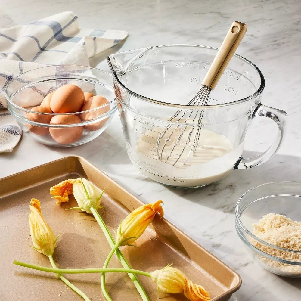 Meja dapur dengan mangkuk telur, mengukur cangkir dengan pengocok dan adonan, bunga labu di atas nampan, dan semangkuk remah roti
