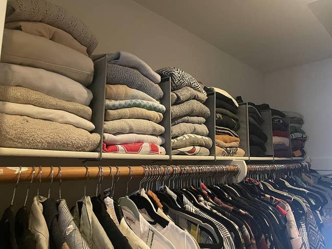 A neatly organized closet with sweaters folded connected shelves and assorted shirts and jackets hanging beneath connected a covering rod