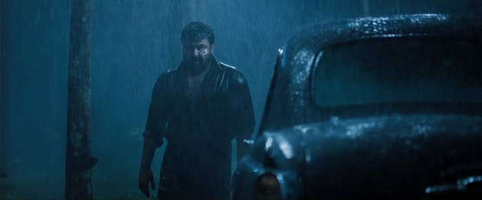 A man stands in the rain, wet shirt clinging to him, beside a vintage car. The scene is dramatic and moody, conveying a sense of tension