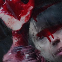 Person with red handprint on glass, looking through it with intense expression. Dark background enhances dramatic feel