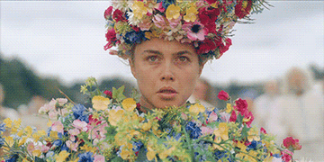 A person wears an elaborate flower crown and outfit, looking focused or intense, in an outdoor setting