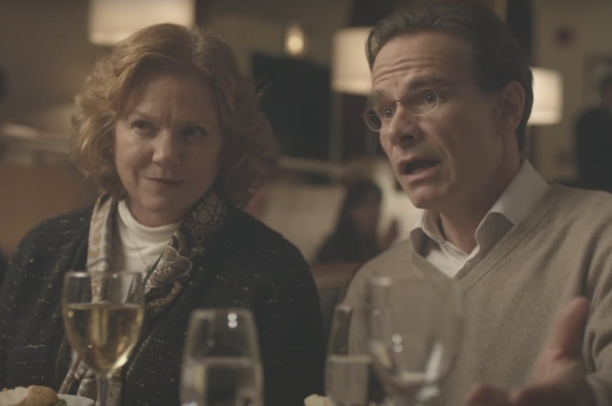 A man and a woman sit at a dinner table with drinks. The woman smiles slightly while the man gestures as he speaks, wearing a sweater over a shirt
