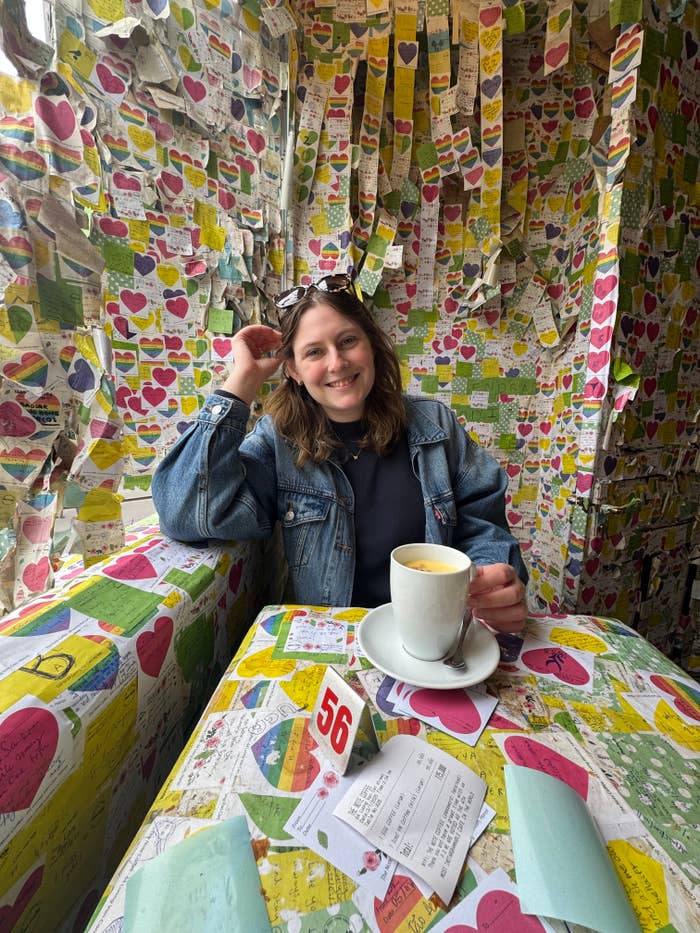 Person in a denim jacket smiling, seated at a cafe table covered with heart notes, holding a coffee. Walls are filled with colorful notes