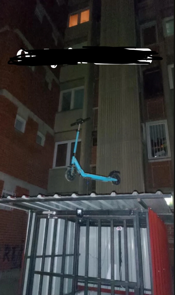 Electric scooter precariously perched on a rooftop structure near a building with windows. Vertical orientation highlights the unusual parking choice