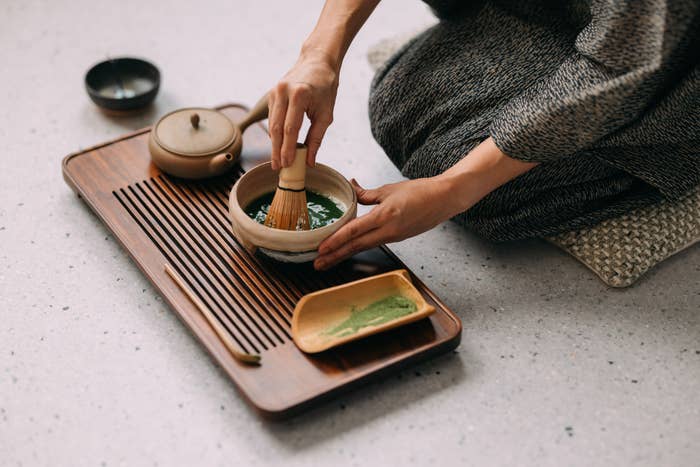 Person preparing matcha tea with a bamboo whisk on a wooden tray, accompanied by a teapot and a small plate of matcha powder