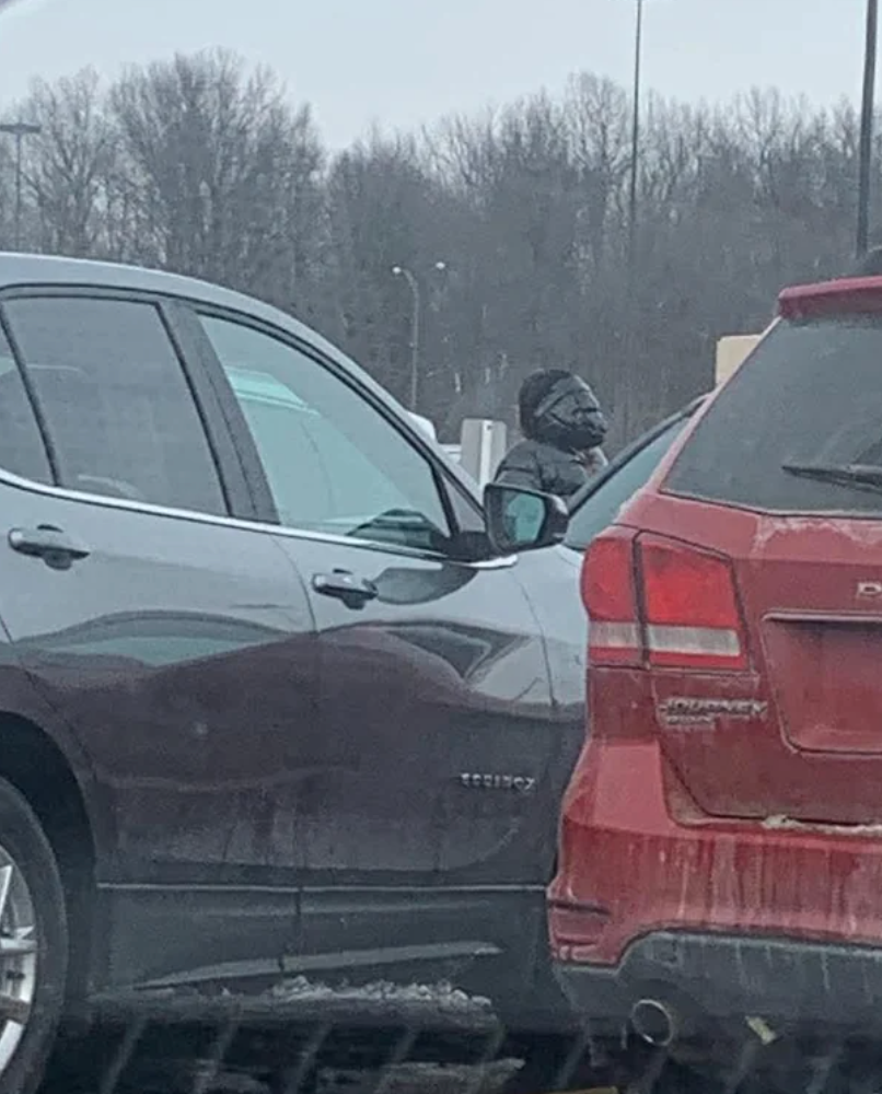 A person wearing a Darth Vader mask is seen in a parking lot between two parked cars