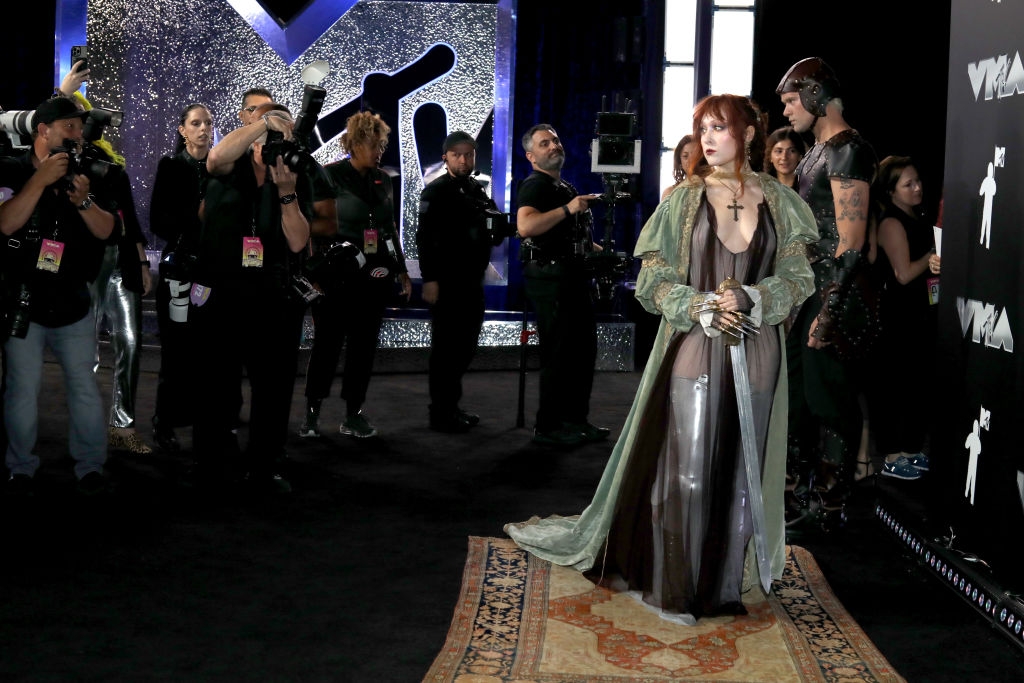 Celebrity on a red carpet in a flowing gown with a cross necklace, surrounded by photographers at a media event