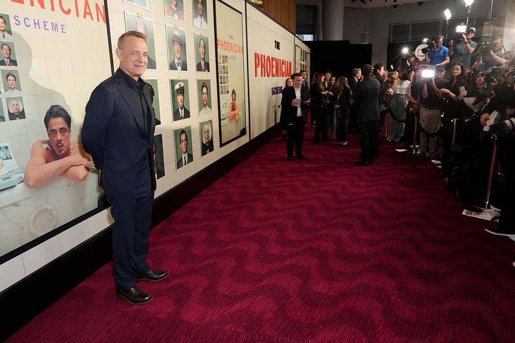 Actor in a suit poses on a red carpet at a film event, with a gallery wall of movie posters and photographers capturing the moment